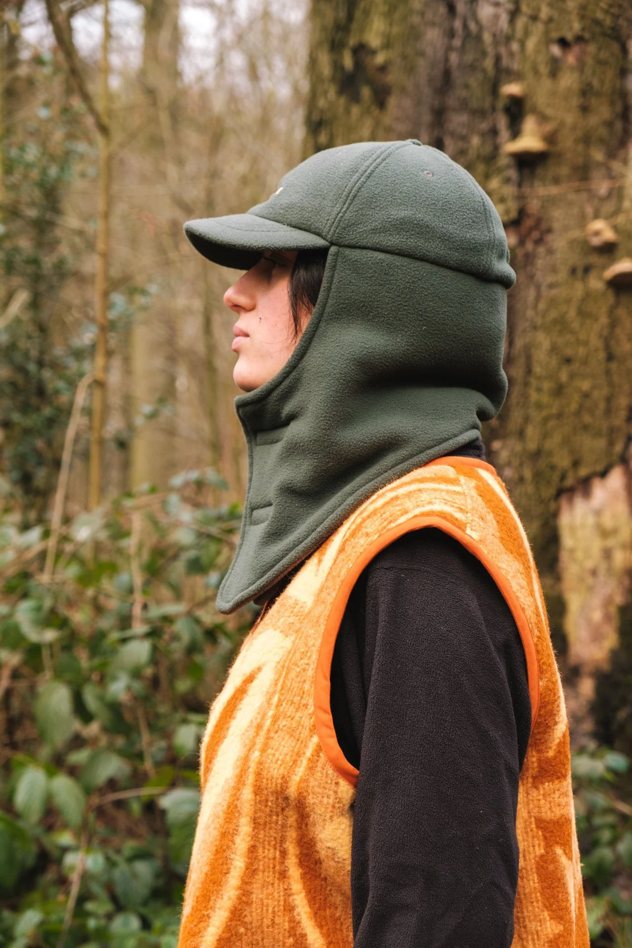 Personne portant une cagoule verte, un gilet orange en peluche et un t-shirt noir, dans une forêt.