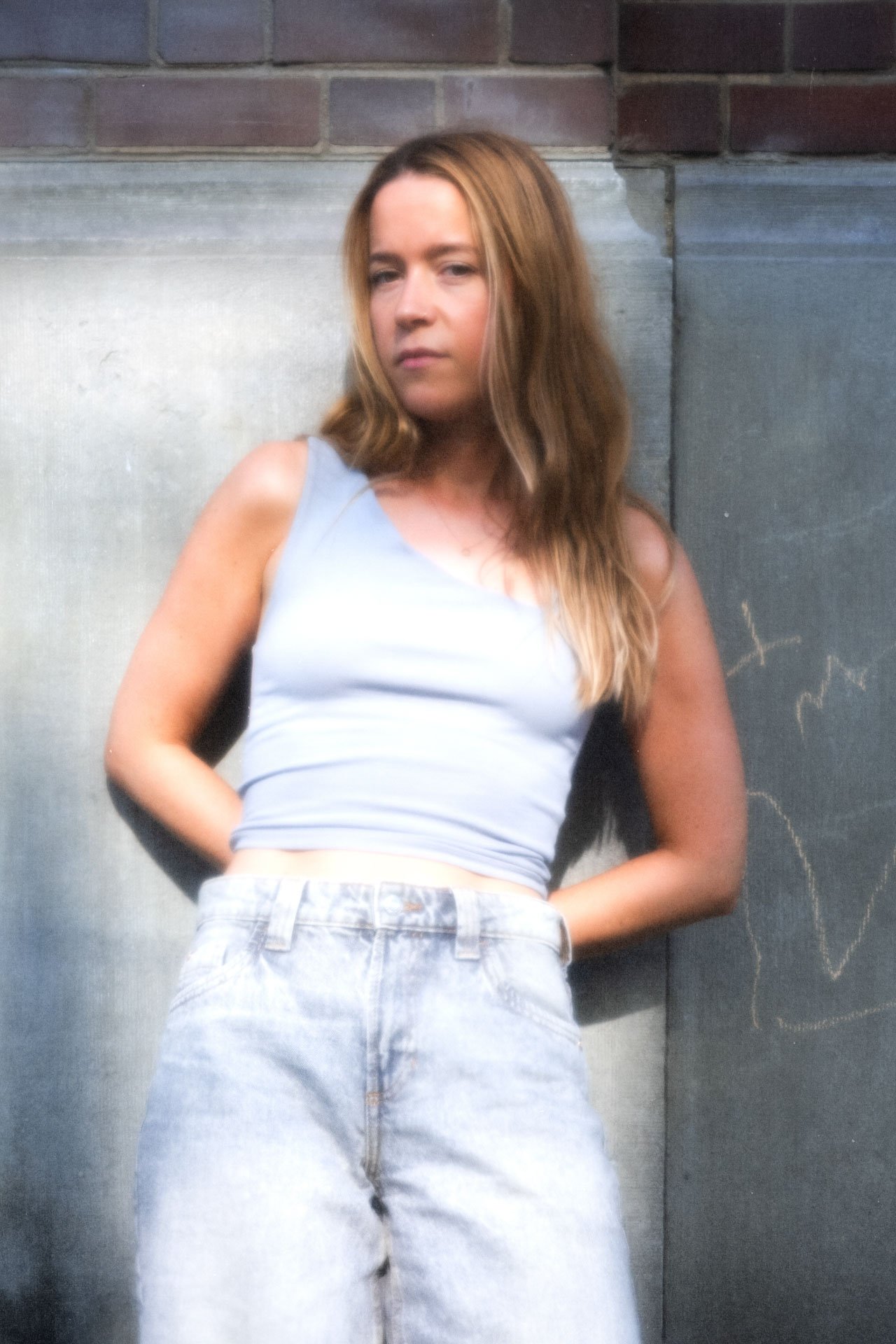 Jeune femme aux cheveux longs et bouclés, portant un haut gris et un jean, debout contre un mur en brique.