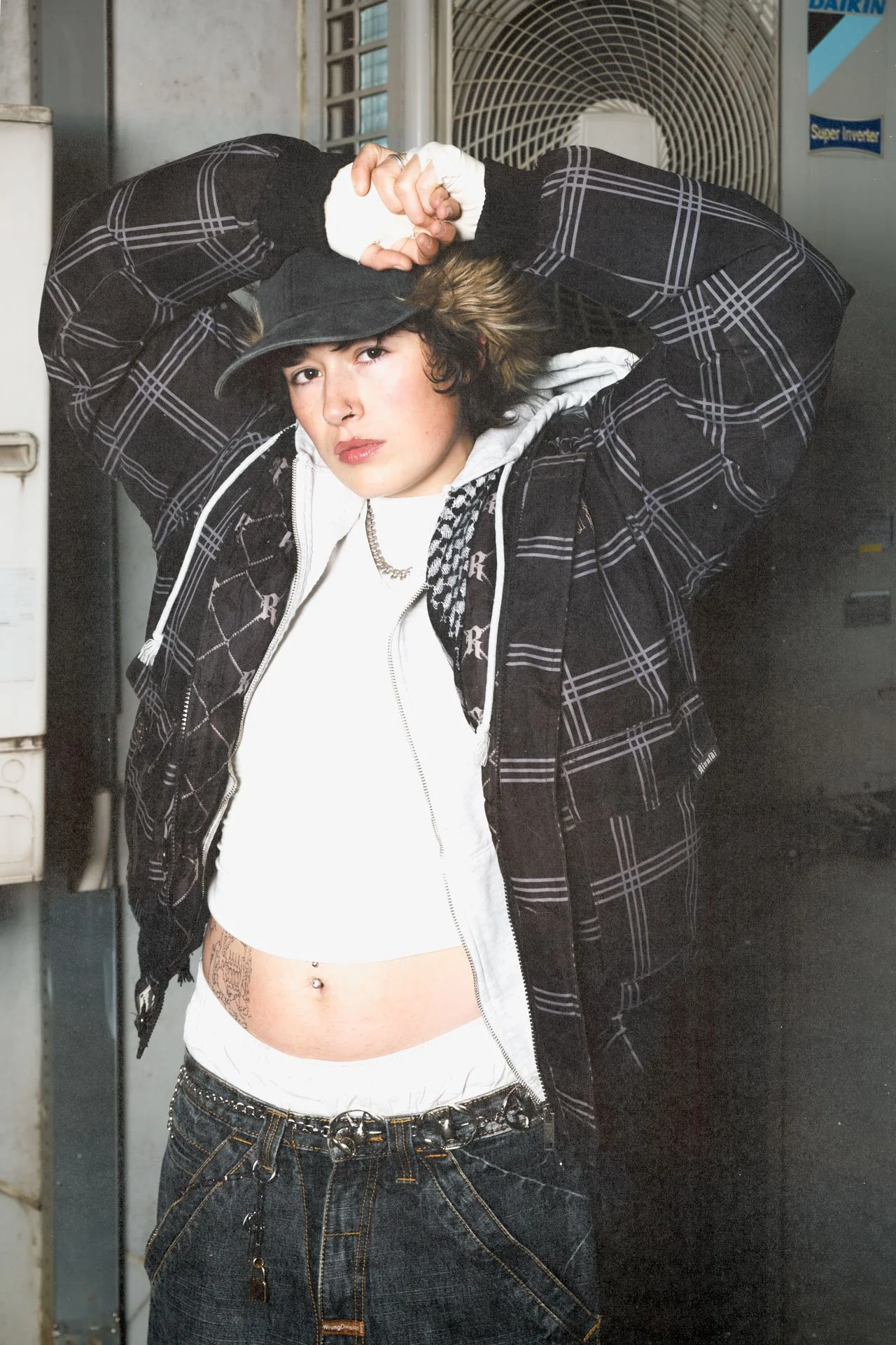Jeune femme posant avec une tenue urbaine devant un mur en béton et une unité de climatisation