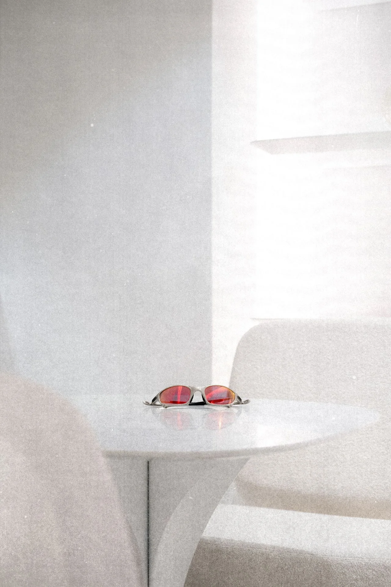 Une paire de lunettes de soleil avec des verres rouges repose sur une étagère blanche, avec des ombres et reflets sur le mur blanc derrière.