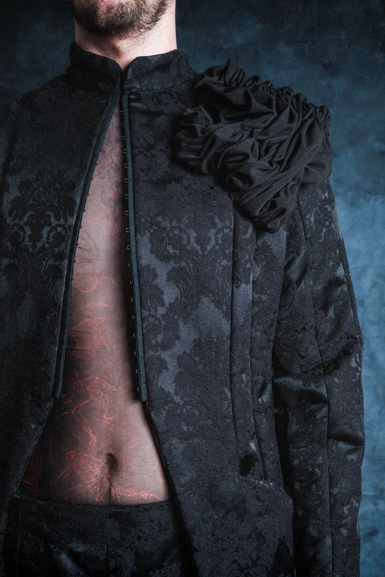 Un homme porte un haut noir avec un motif en relief et une grande broderie de tissu dans les épaules, avec un tissu transparent avec un motif rouge visible sous le haut. Le fond est sombre et huileux.