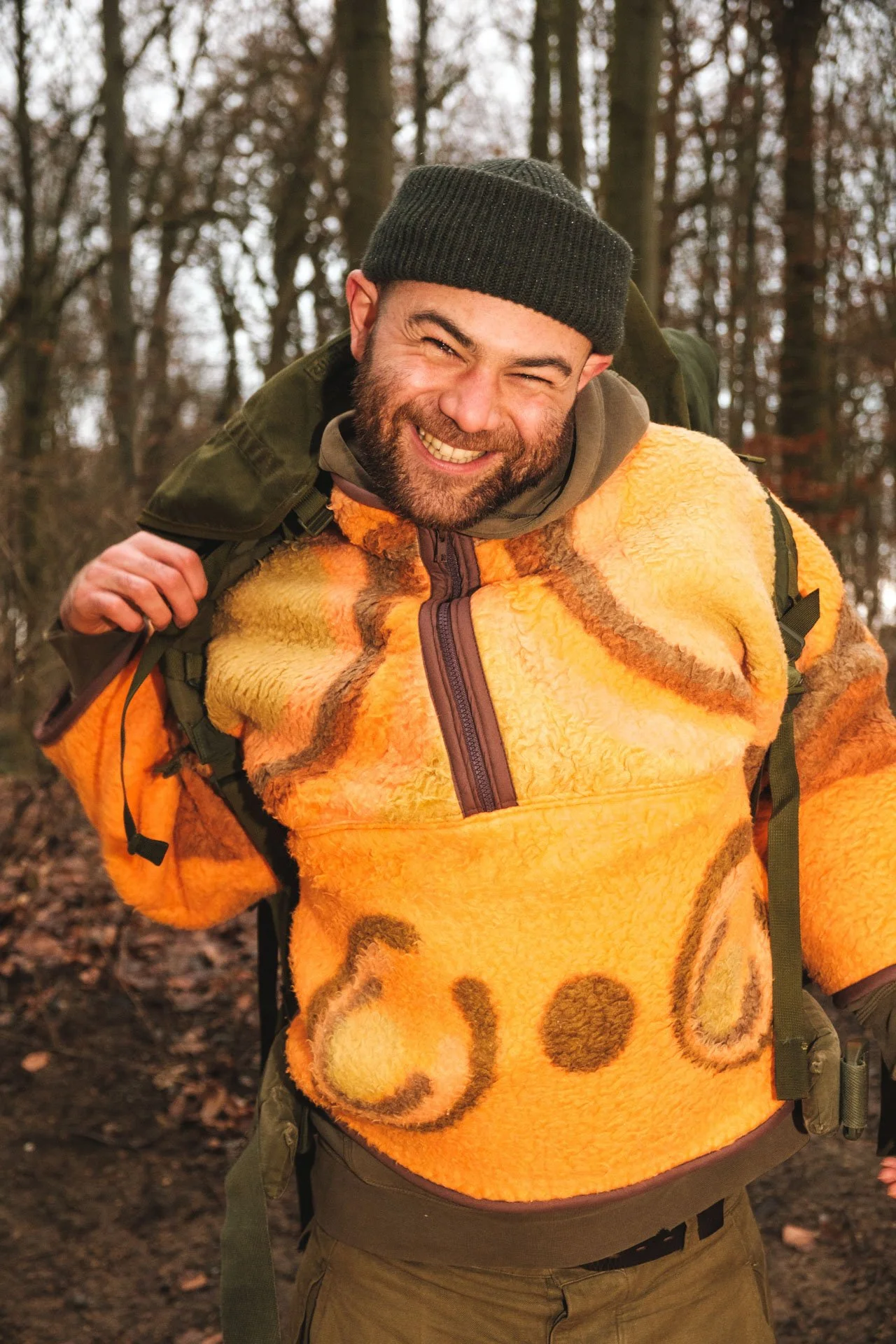 Un homme souriant en plein air dans une forêt, portant un bonnet gris, un pull orange chaud avec un motif de cercles, et un sac à dos vert.