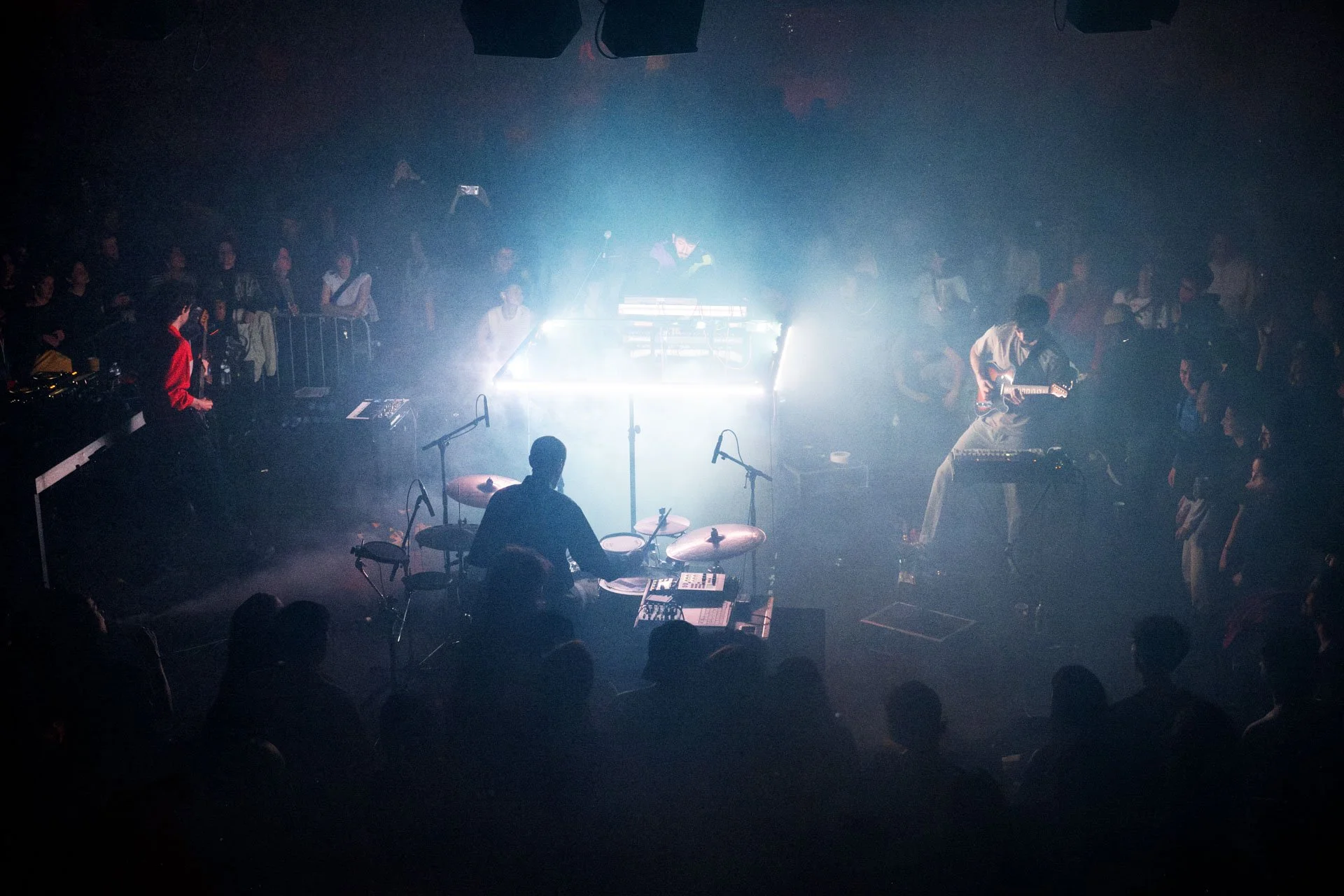 Une scène de concert dans une salle sombre avec un groupe de musiciens jouant sous des lumières faibles. Il y a un batteur au premier plan, un guitariste à droite, un clavier à gauche, et plusieurs personnes dans le public qui regardent la scène.