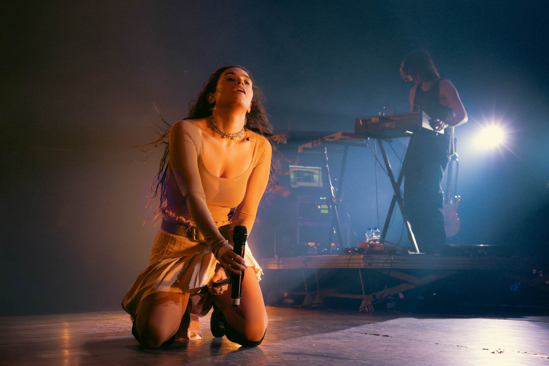 Une femme qui chante en position inclinée, tenant un microphone, avec une personne en arrière-plan manipulant des équipements électroniques sur scène, sous des lumières de spectacle.