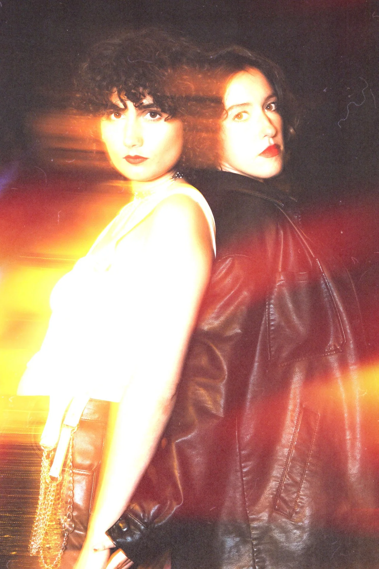 Deux femmes posant dos à dos, l'une avec des cheveux bouclés et l'autre avec des cheveux raides, vêtues de tenues foncées et de vêtements en cuir, sous une lumière vive et colorée.