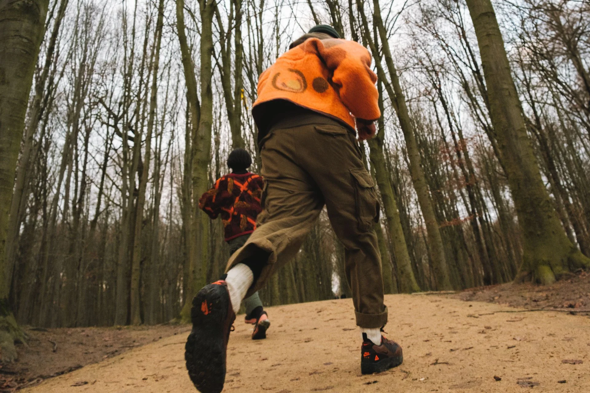 Deux enfants qui courent dans un bois en automne, vu d'en dessous, avec des arbres dénudés et un sol en terre.