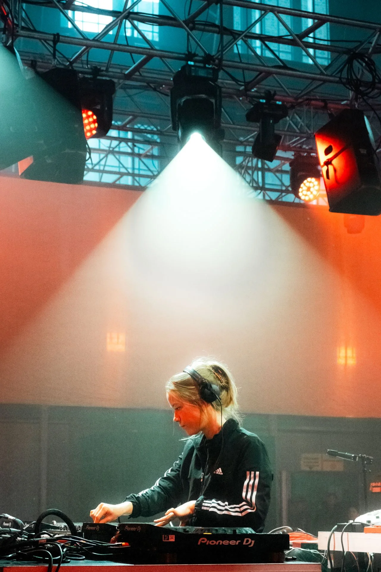 Une femme DJ avec des écouteurs, utilisant un équipement de musique Pioneer DJ, dans un espace avec éclairage de scène rouge et verte, en train de mixer.
