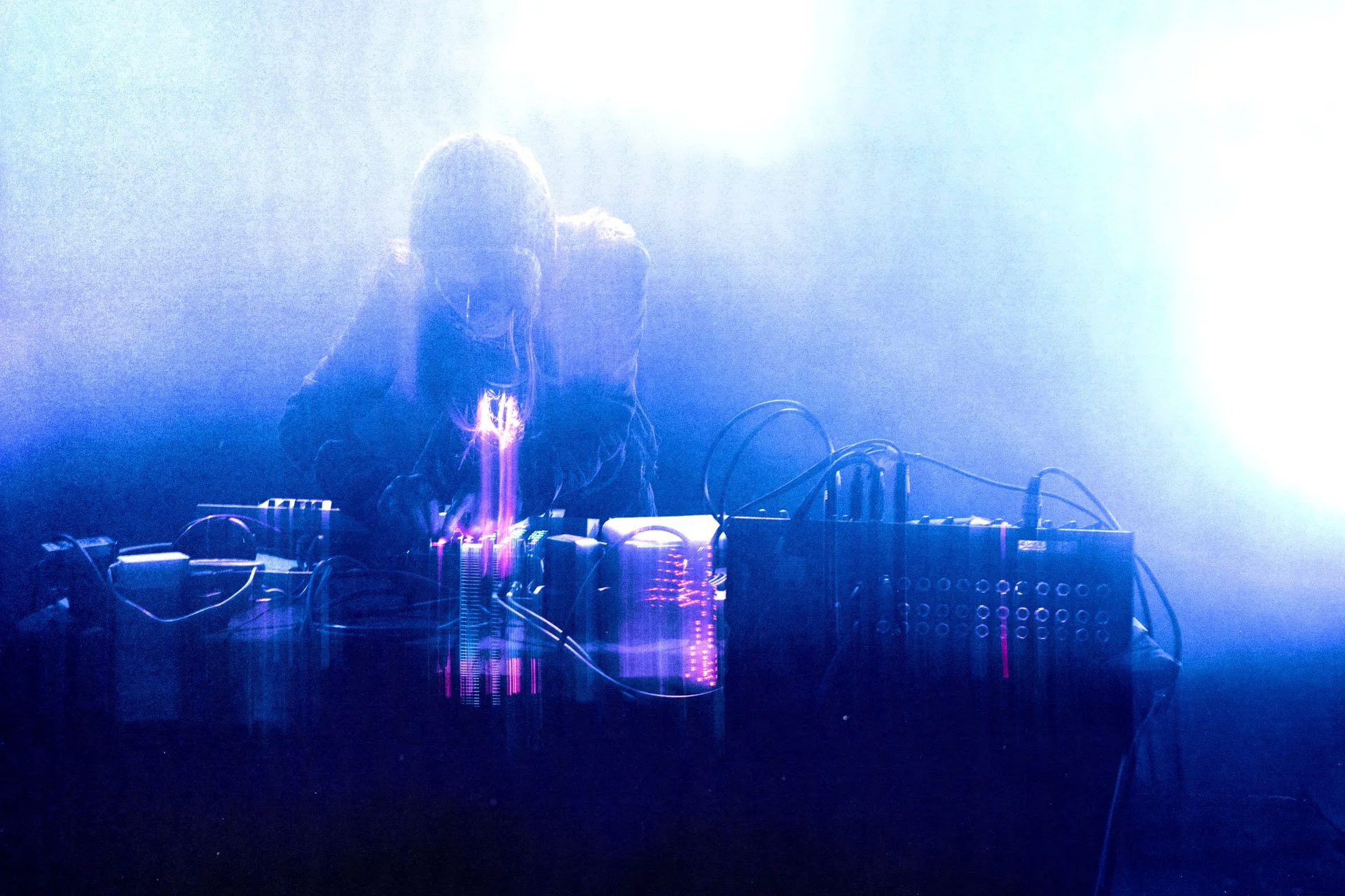 DJ en train de mixer sur scène, entouré de câbles et d'équipements, avec une lumière bleue et une ambiance mystérieuse.