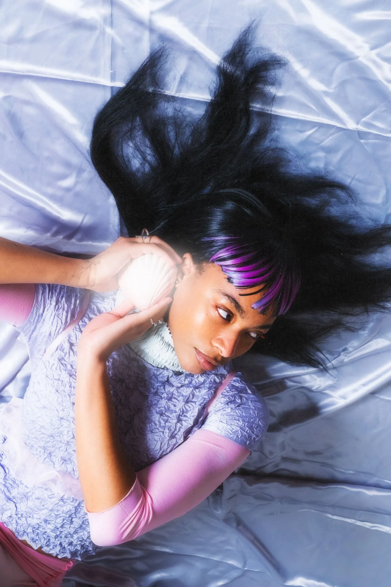 Une jeune femme avec des cheveux noirs et violets, portant un haut rose et blanc, repose sur un lit en satin blanc, tenant une coquille de mer près de l'oreille.