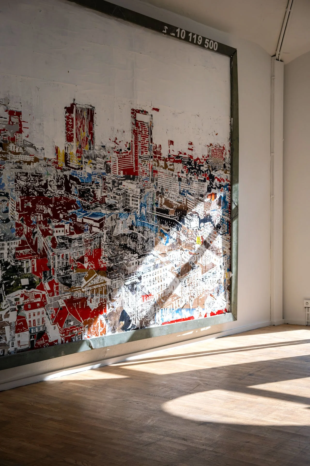Mur avec une affiche représentant une ville en détails colorés, encadrée par un cadre en métal, au sol un parquet en bois, et des rayons de lumière naturelle qui tombent au sol.