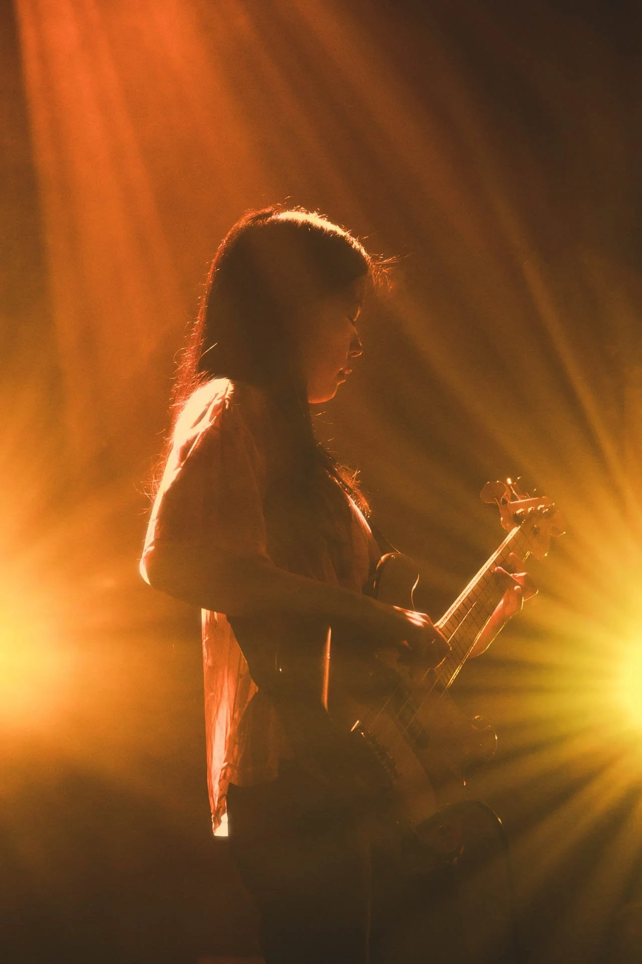 Jeune femme jouant de la guitare électrique sur scène sous un éclairage orange et jaune.