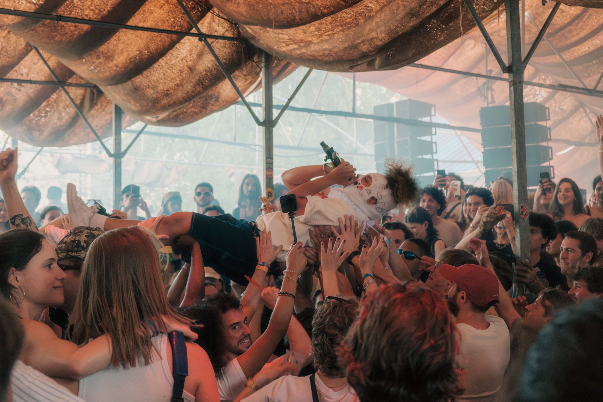 Groupe de personnes lors d'un concert ou festival en intérieur, une personne est portée sur la foule, d'autres tiennent des appareils photos ou enregistrent. L'atmosphère est festive et animée.