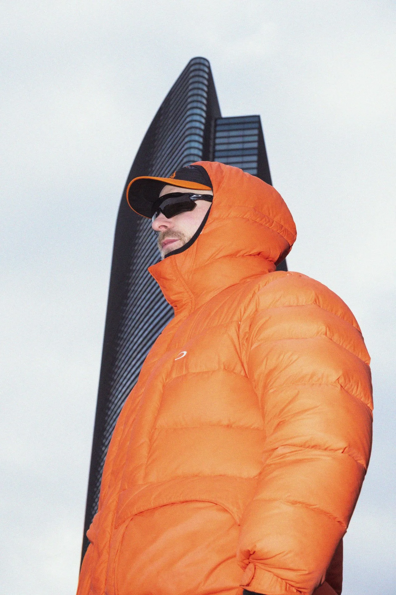 Un homme portant un manteau orange, des lunettes de soleil et une casquette, avec un bâtiment en forme de voile derrière lui, en plein air sous un ciel gris.