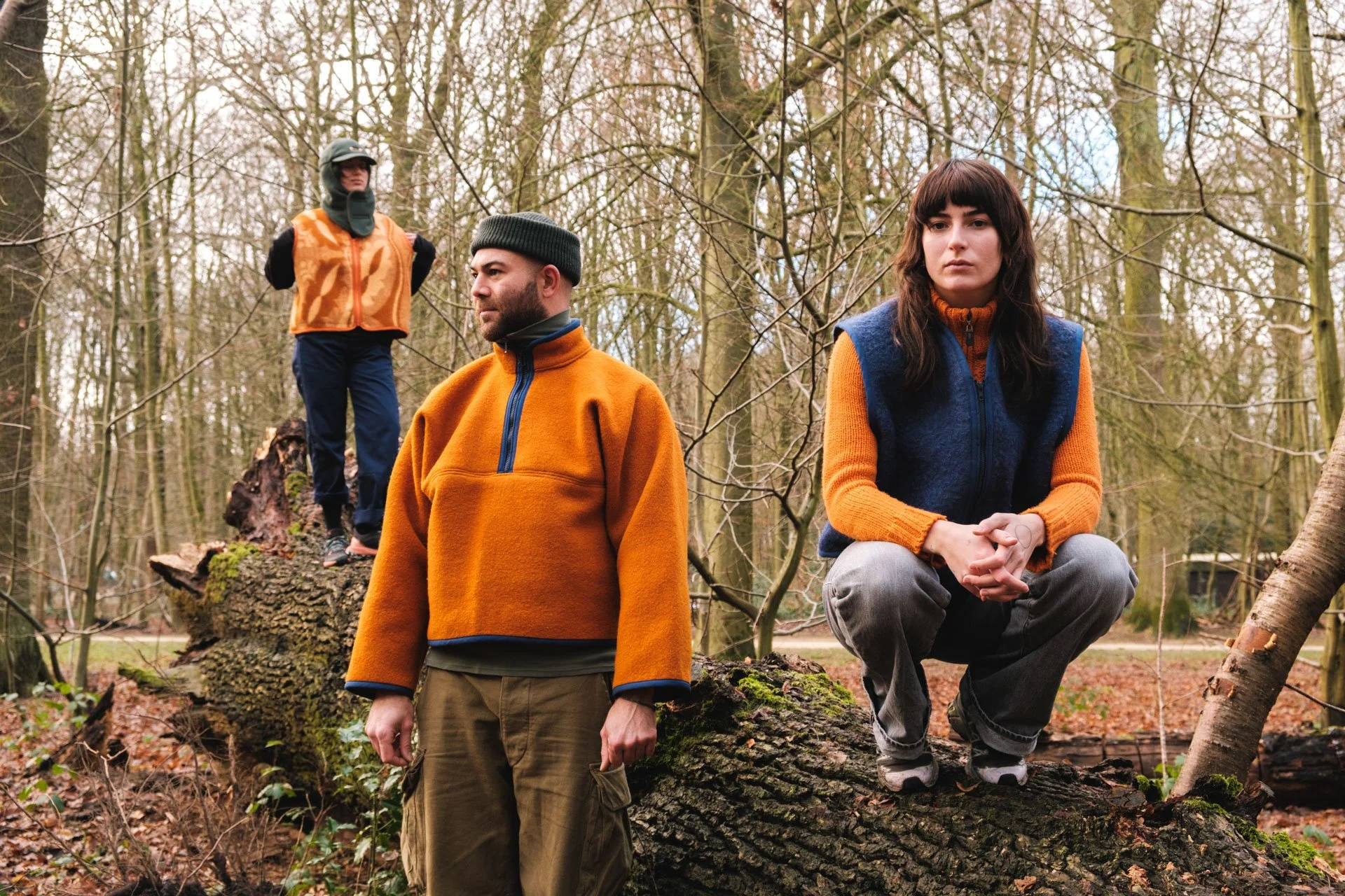 Trois personnes dans une forêt, deux debout devant un gros tronc d'arbre couché et une assise sur une branche, vêtements d'extérieur colorés, paysage forestier en arrière-plan.