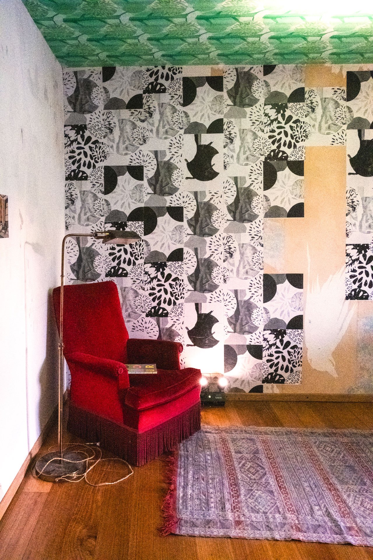 Un coin salon avec un fauteuil rouge en velours, une lampe sur pied, un tapis traditionnel sur un parquet en bois, et un mur avec un papier peint à motifs en noir et blanc.