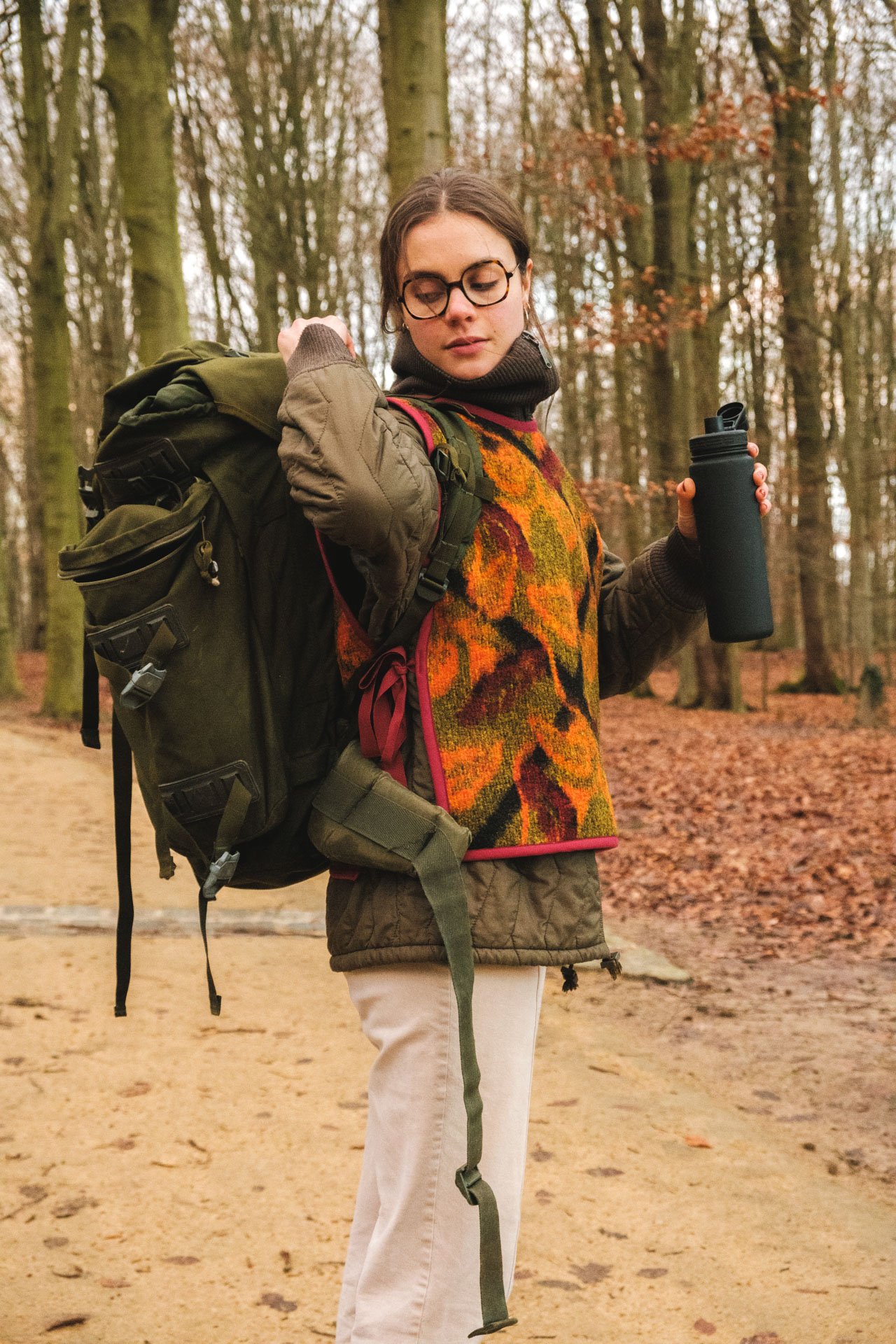 Une femme portant un sac à dos orange et vert, un sweat-shirt coloré, des lunettes, et tenant une bouteille d'eau, marche dans une forêt d'automne.