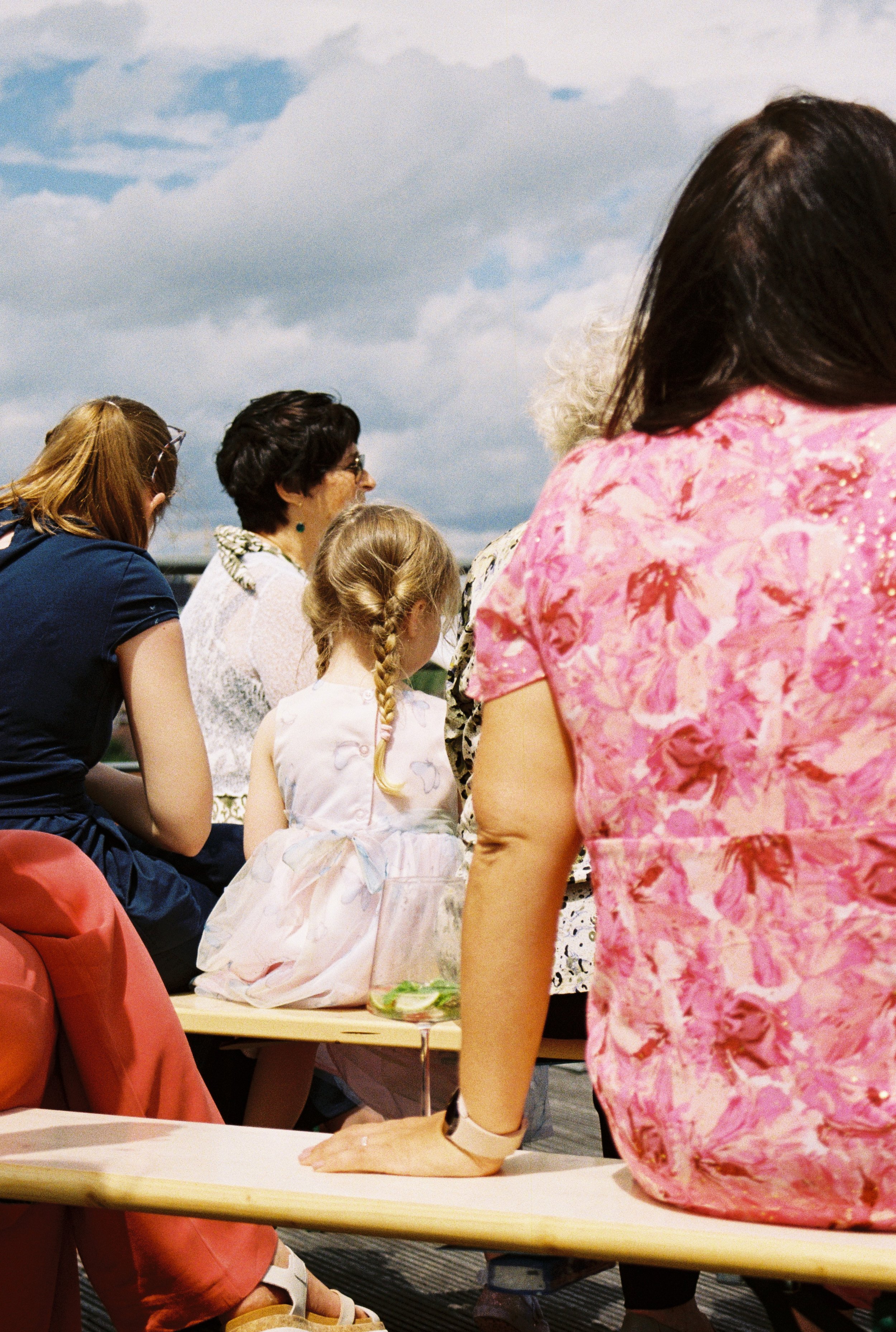 Groupe de personnes assises en plein air lors d'un rassemblement, avec un ciel nuageux en arrière-plan, principalement visibles la partie supérieure du corps des femmes et un enfant avec des cheveux blonds attachés.