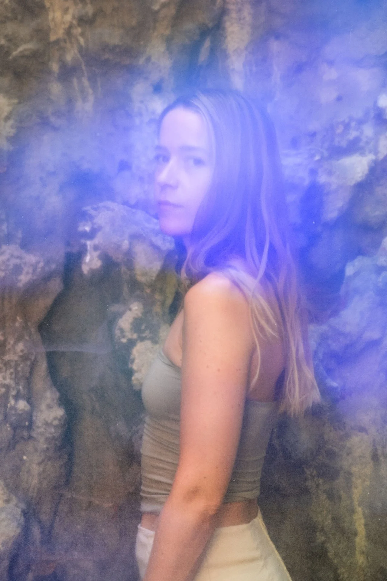 Une jeune femme avec de longs cheveux bouclés, portant un débardeur gris, assise contre une paroi rocheuse dans une grotte, regardant vers l'objectif.