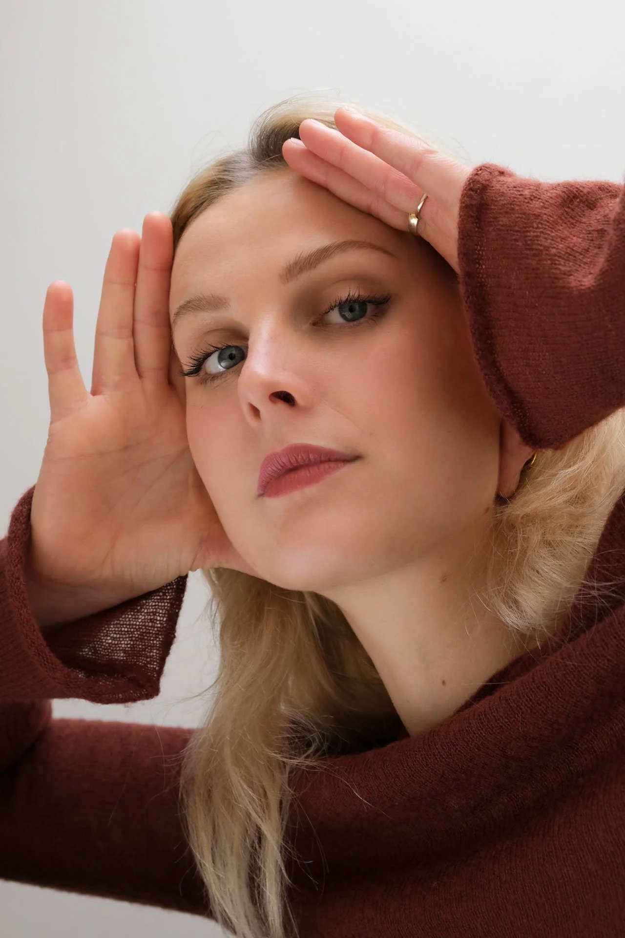 Une femme blonde aux yeux bleus pose avec ses mains près de son visage, portant un pull marron et un fond blanc.