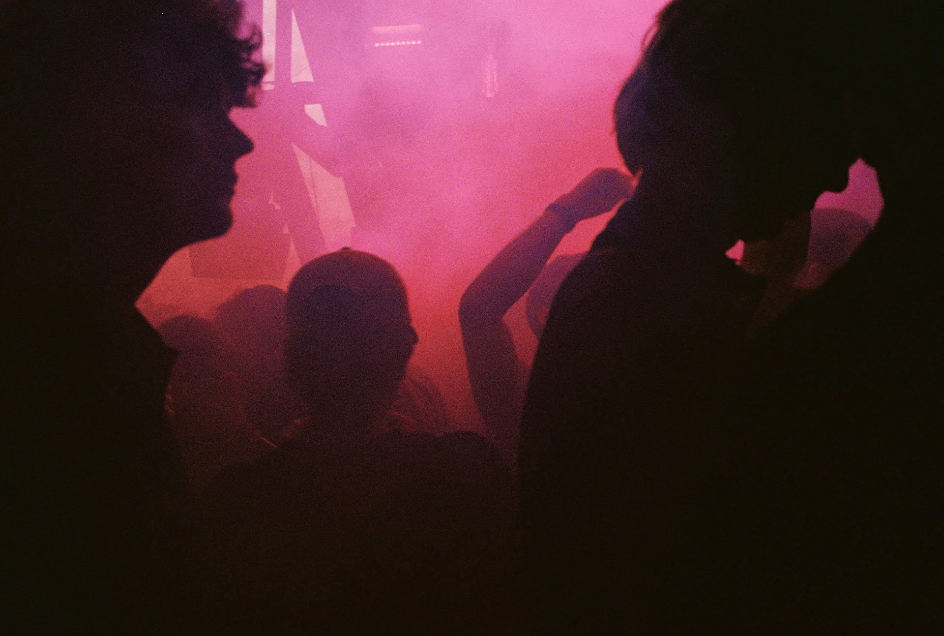 Silhouettes de personnes dans une ambiance sombre avec un éclairage rose et violet, évoquant une scène de fête ou de club