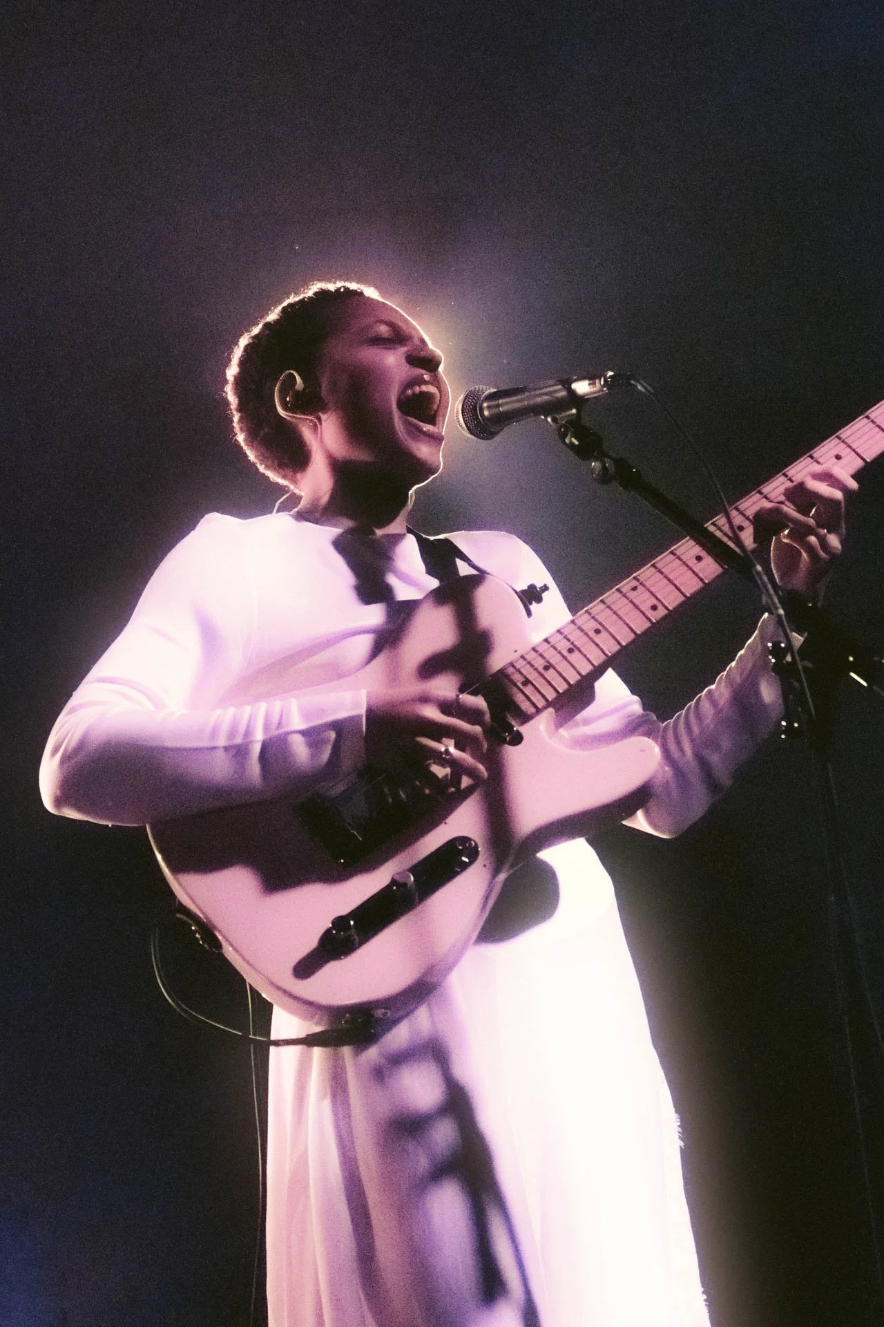 Une femme chante en jouant de la guitare électrique lors d'une performance musicale, éclairée par une lumière douce et sombre autour.