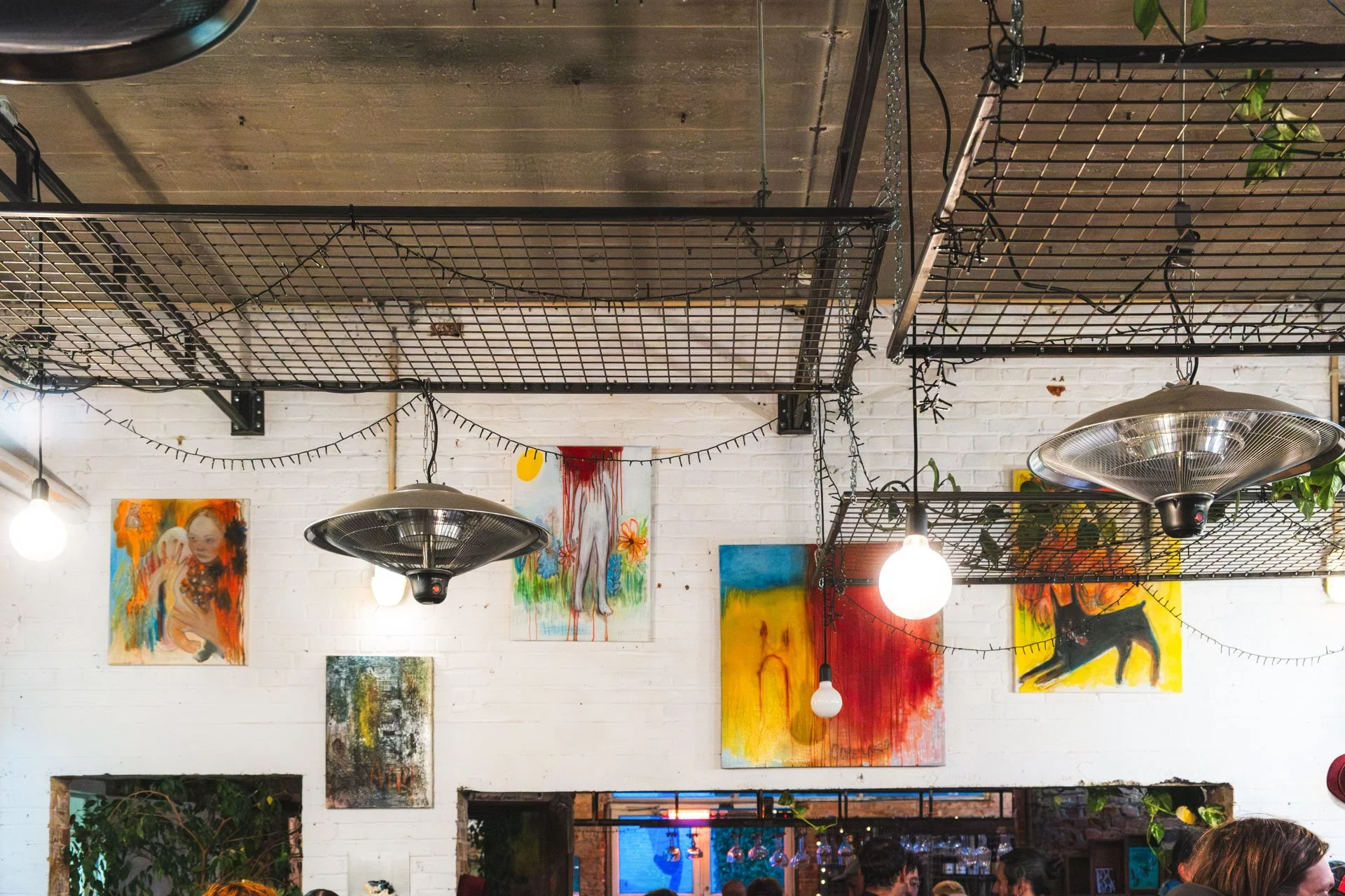 Une vue intérieure d'un café ou galerie d'art avec éclairage suspendu, peintures colorées sur mur, décor végétal et plafond en bois