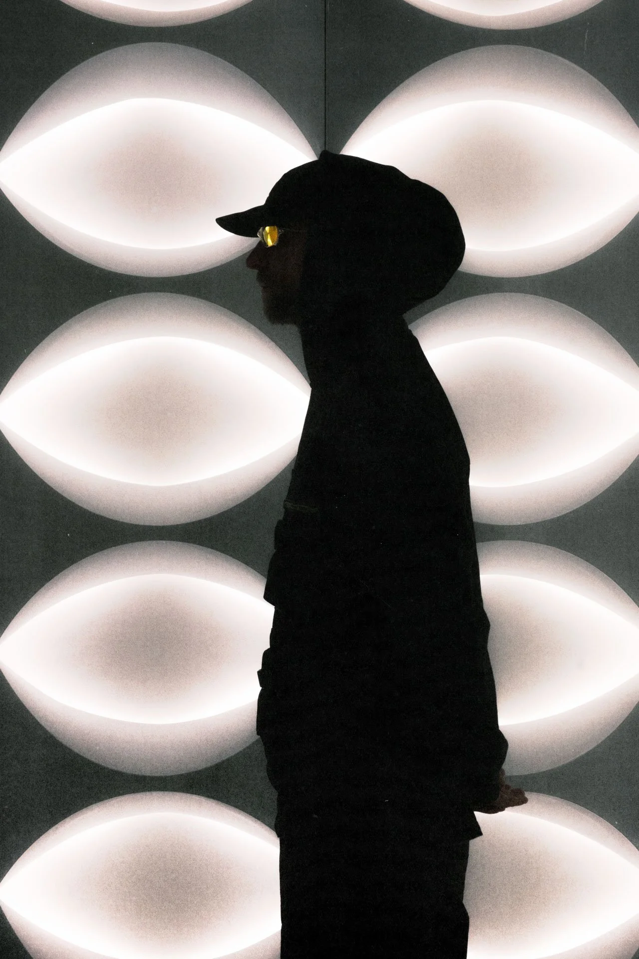 Silhouette d'une personne portant une casquette, avec des lunettes de soleil, devant un mur de lumières blanches arrondies.