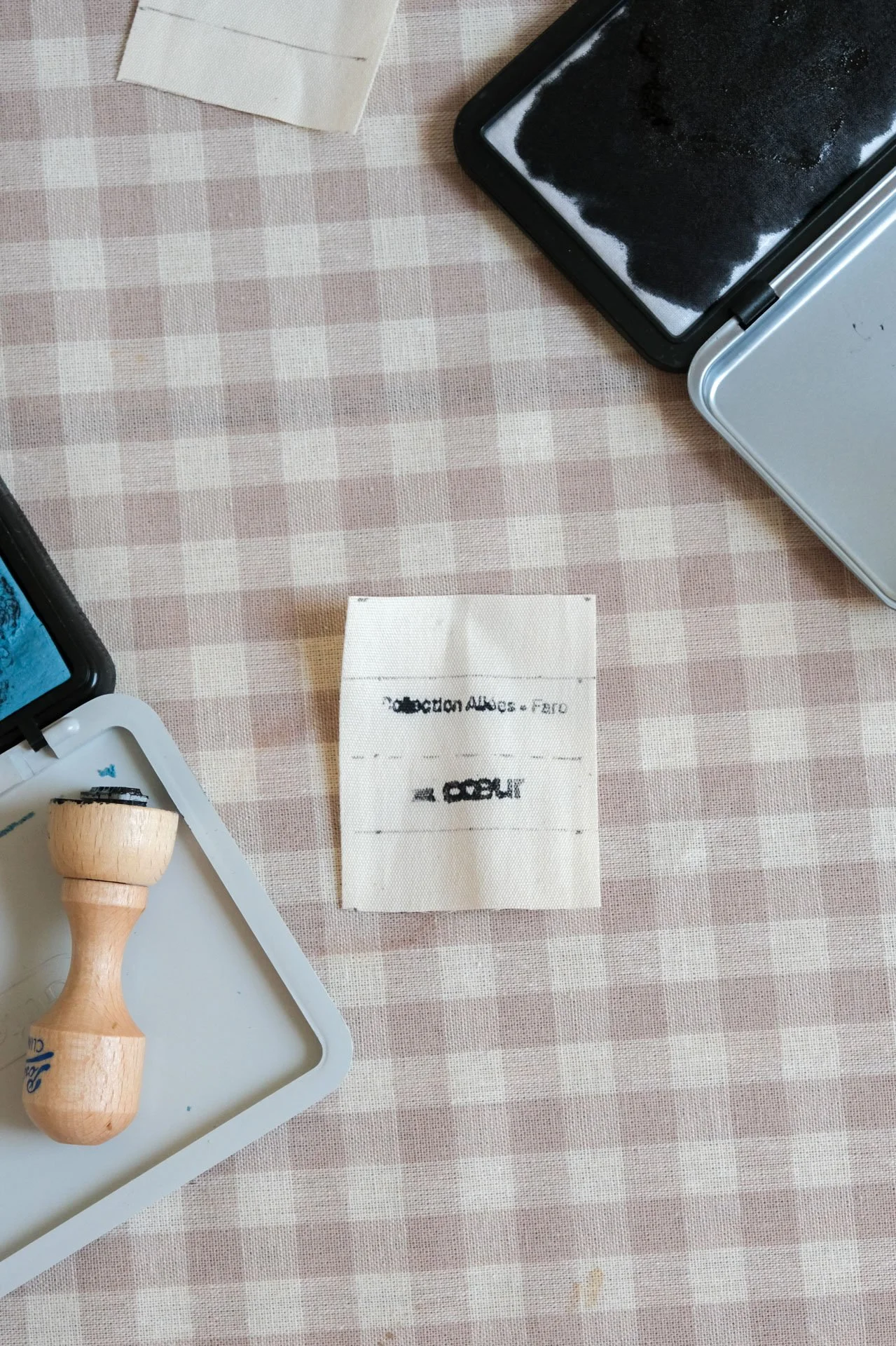 Une table avec un motif à carreaux rose et blanc, contenant divers objets de bureau et artisanaux, y compris un rouleau de tampon en bois, un tampon encreur noir, une petite étagère en plastique avec un tampon, un carré d'encre déposée, une tige en p