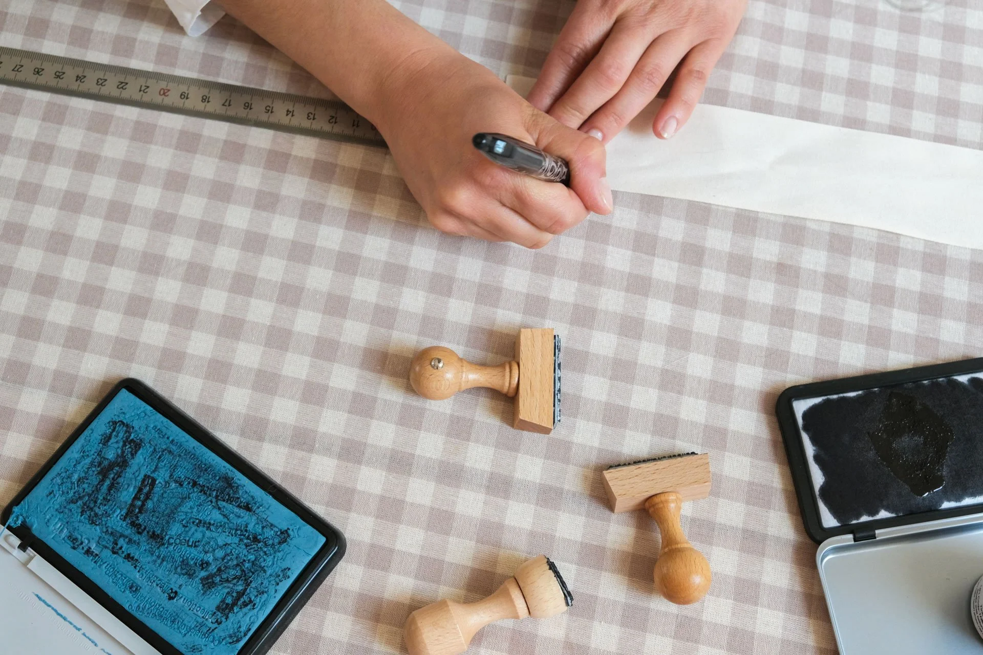 Une personne écrivant avec un stylo noir sur une feuille de papier posée sur une table à carreaux. Il y a une règle en métal, des tampons en bois, un smartphone, et une boîte avec de l'encre noire.