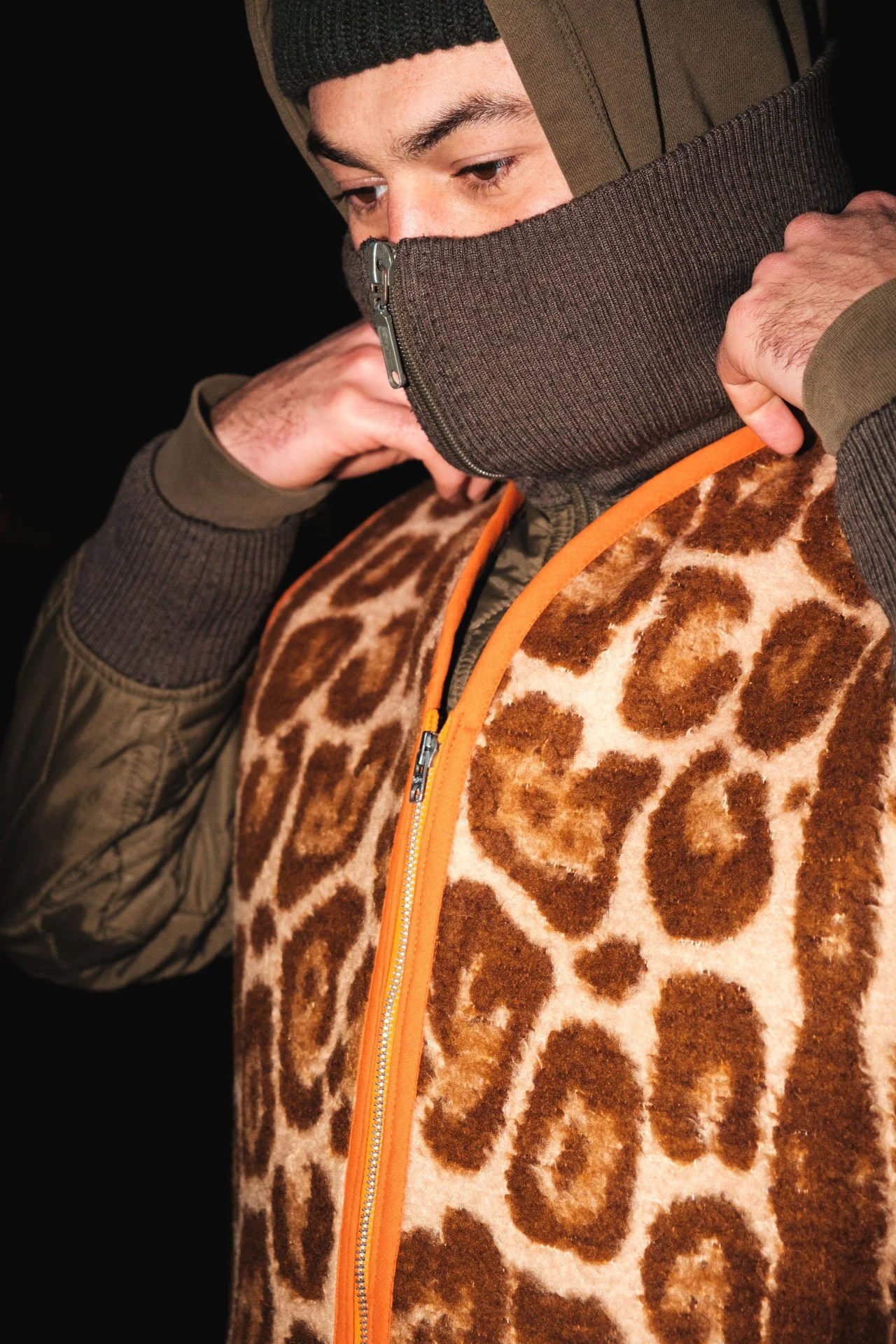 Un homme porte un pull avec un motif de léopard et un vestiaire vert avec un détail orange sur la fermeture éclair. Il porte également un bonnet noir et un cache-cou marron foncé.