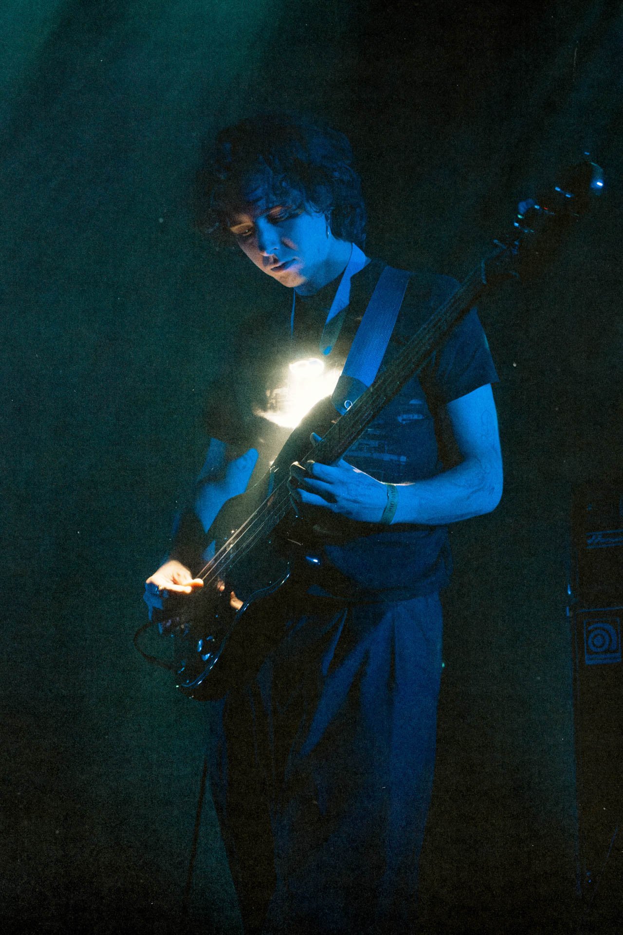 Un guitariste joue dans une scène sombre, éclairé par une lumière douce, portant un T-shirt à motifs, avec un instrument de musique électrique.