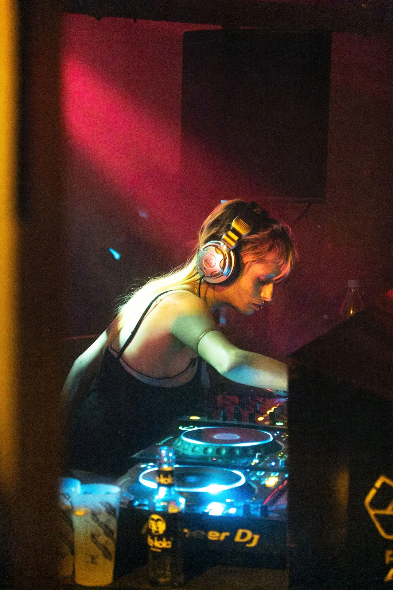 Une femme DJ avec des écouteurs, utilisant un contrôleur DJ dans une discothèque sombre avec des lumières colorées.