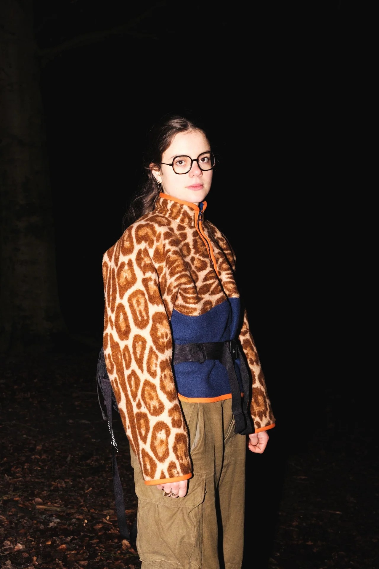 Jeune femme portant un manteau à motif léopard, des lunettes, et des vêtements décontractés, photo prise la nuit dans un environnement nocturne.
