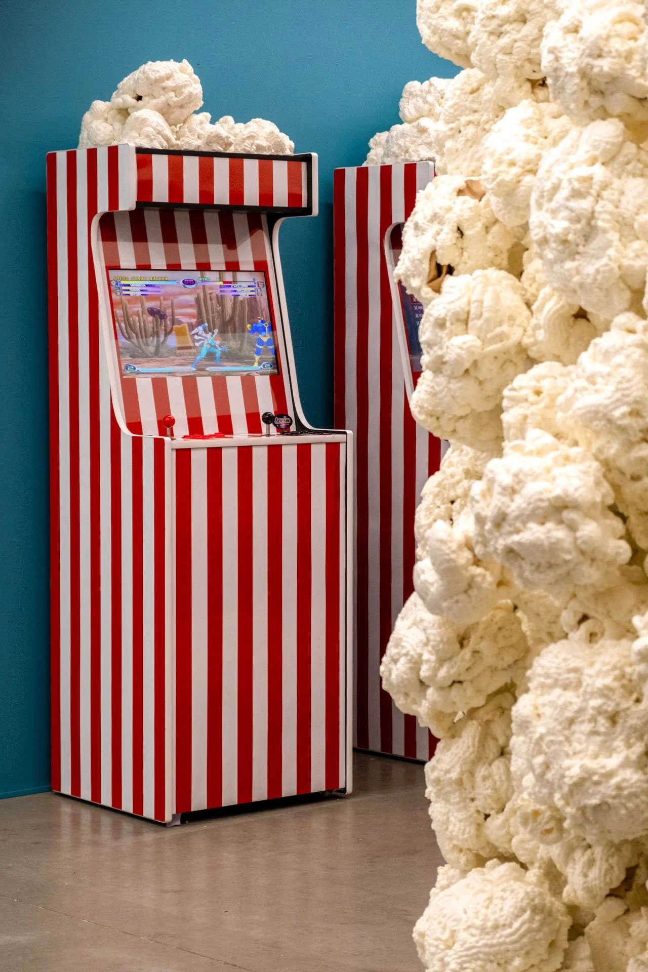 Une borne d'arcade rouge et blanc avec un écran de jeu vidéo, située dans une pièce avec un mur bleu, et des morceaux de popcorn géants devant.