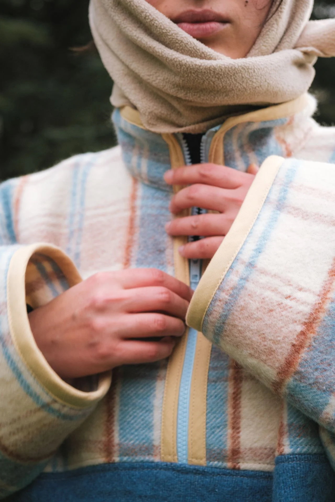 Personne portant une veste colorée à capuche et un foulard beige, tenant son col avec les mains.