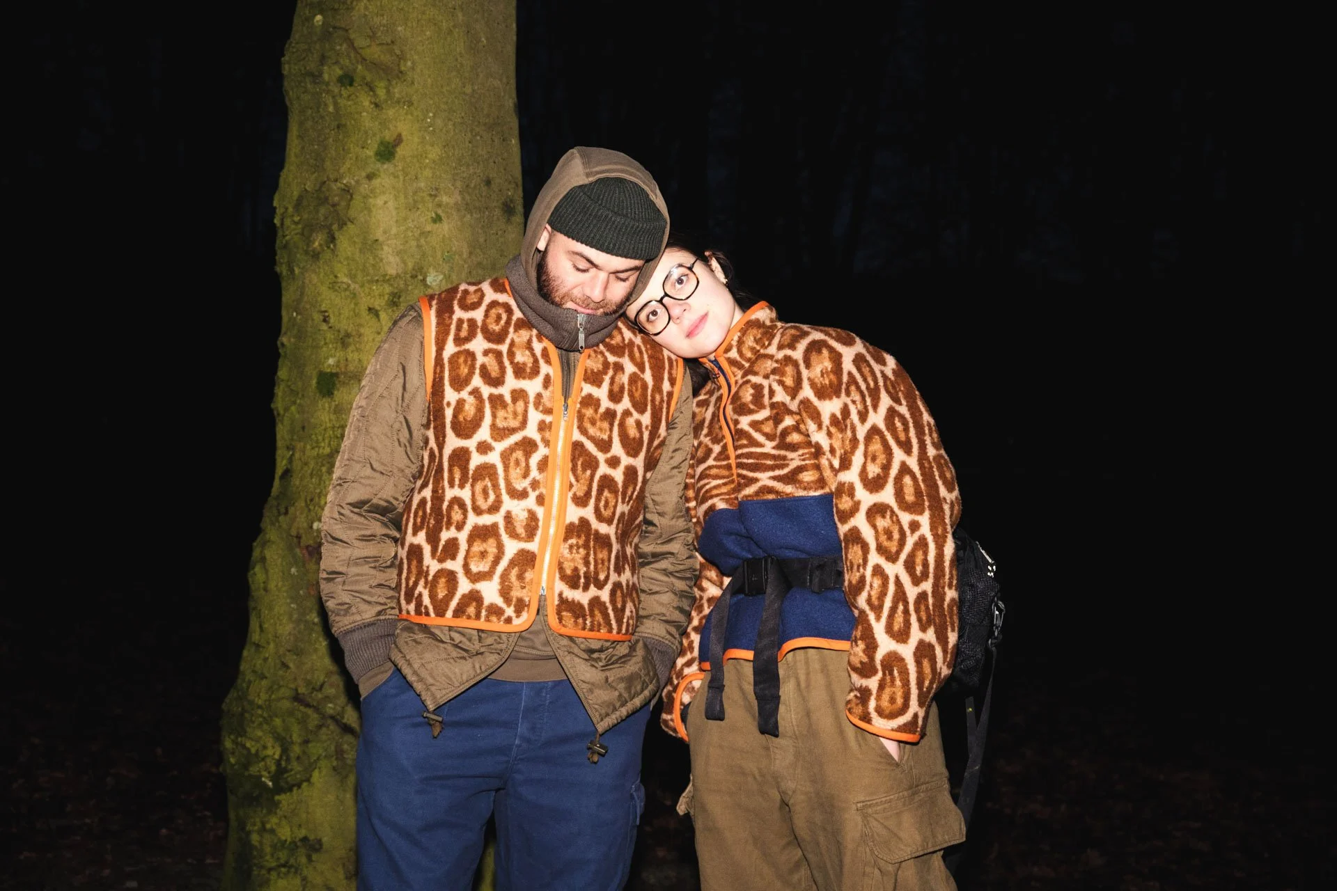 Deux personnes portant des vestes à motif léopard se tiennent proches l'une de l'autre devant un arbre dans une forêt sombre.