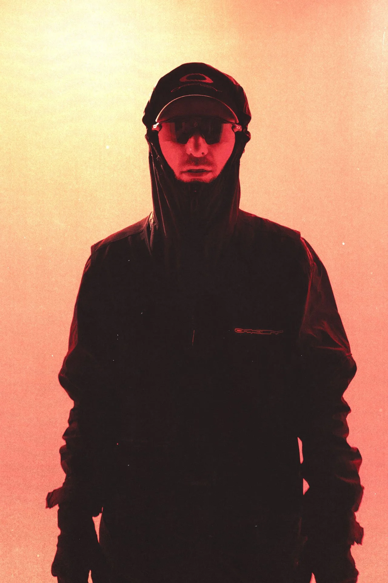 Un homme en manteau noir, lunettes de soleil et casquette, regardant frontalement, avec un fond rouge orangé.