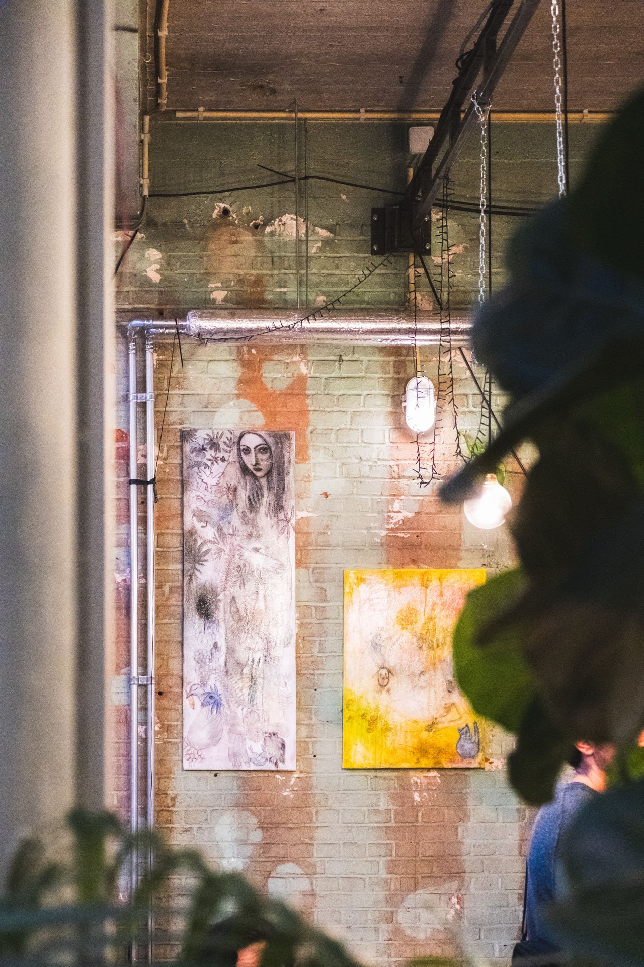 Mur en briques avec deux œuvres d'art suspendues, éclairage suspendu, et présence de personnes floues dans le premier plan.