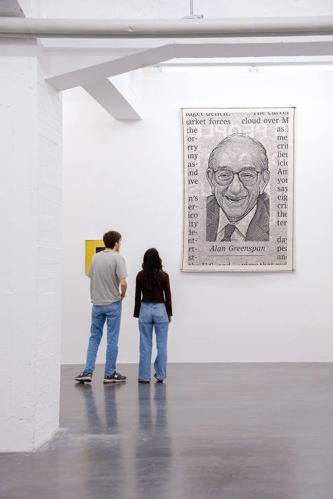 Deux personnes regardent une œuvre d'art dans une galerie blanche, une grande affiche avec le portrait en noir et blanc d'un homme portant des lunettes et une cravate, signé Alan Greenspan.