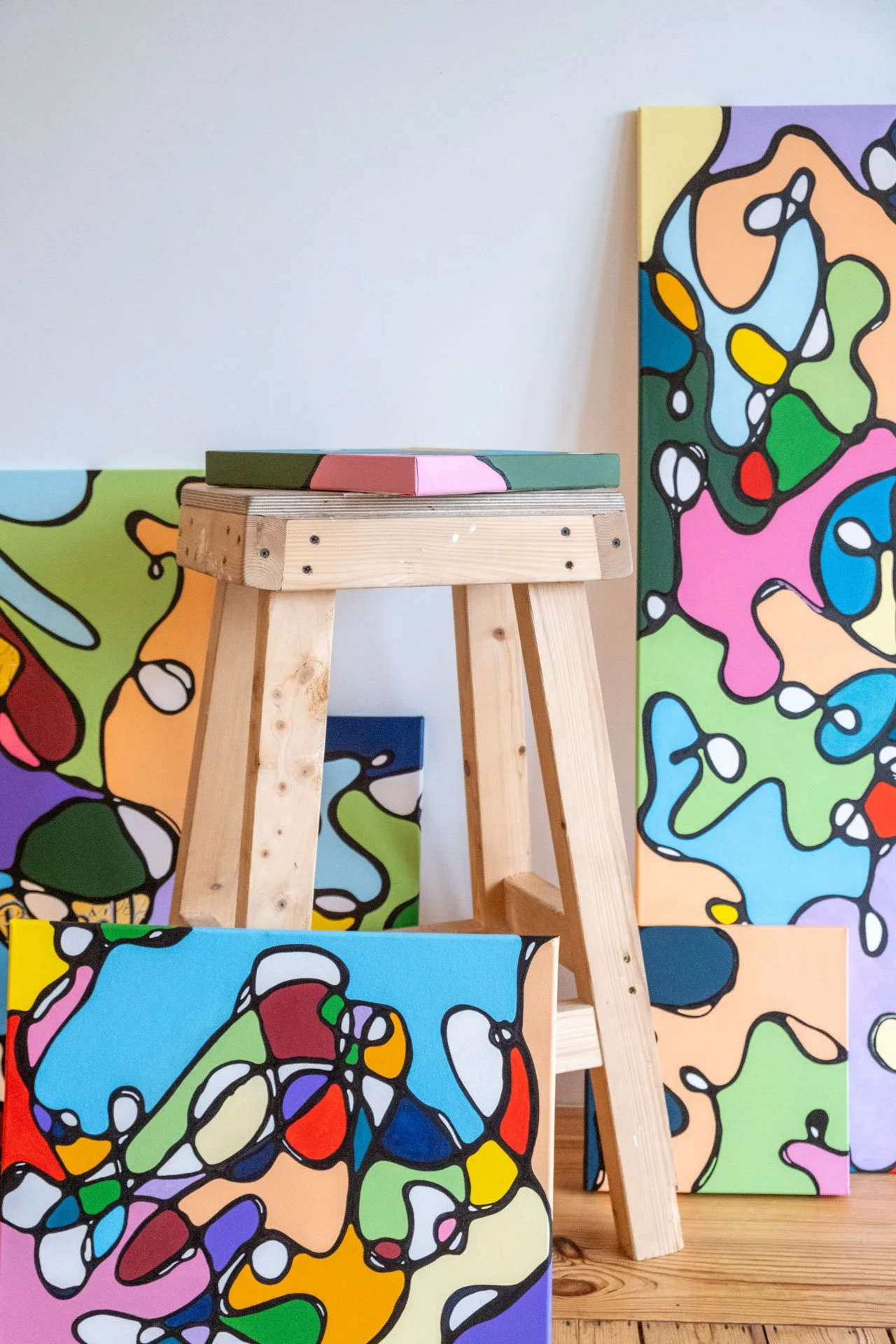 Peintures abstraites colorées avec des formes organiques et des contours noirs, disposées autour d'un trépied en bois dans une galerie d'art.
