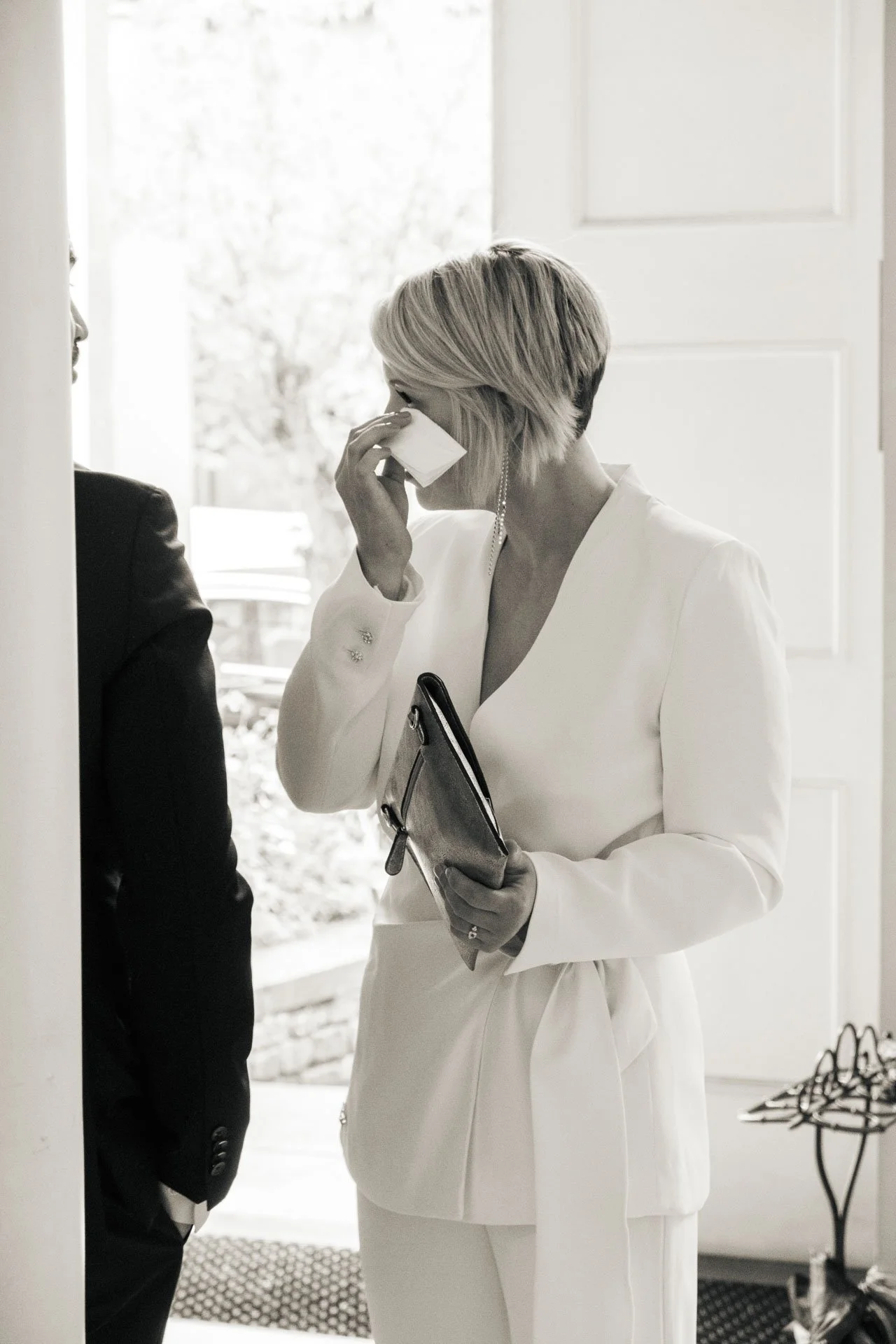 Femme en tenue blanche, tenant un foulard ou un portefeuille et utilisant un mouchoir pour se couvrir le visage, se tenant près d'une porte d'entrée dans une maison.