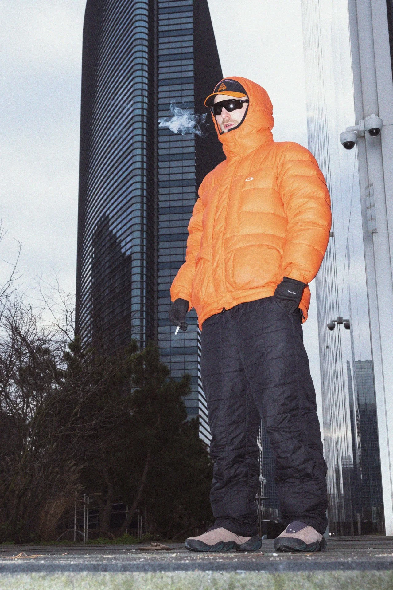 Un homme portant une veste orange, un pantalon noir, des chaussures de sport, des lunettes de soleil et un chapeau orange, fumant une cigarette dans un environnement urbain avec de grands immeubles en arrière-plan.