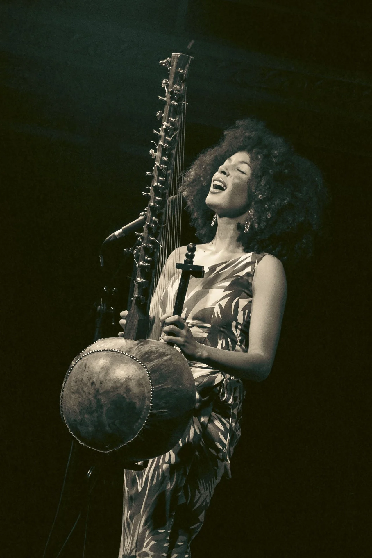 Une femme noire avec de longues boucles cheveux joue d'un instrument de percussion et d'une harpe, en chantant, portant une robe rayée.