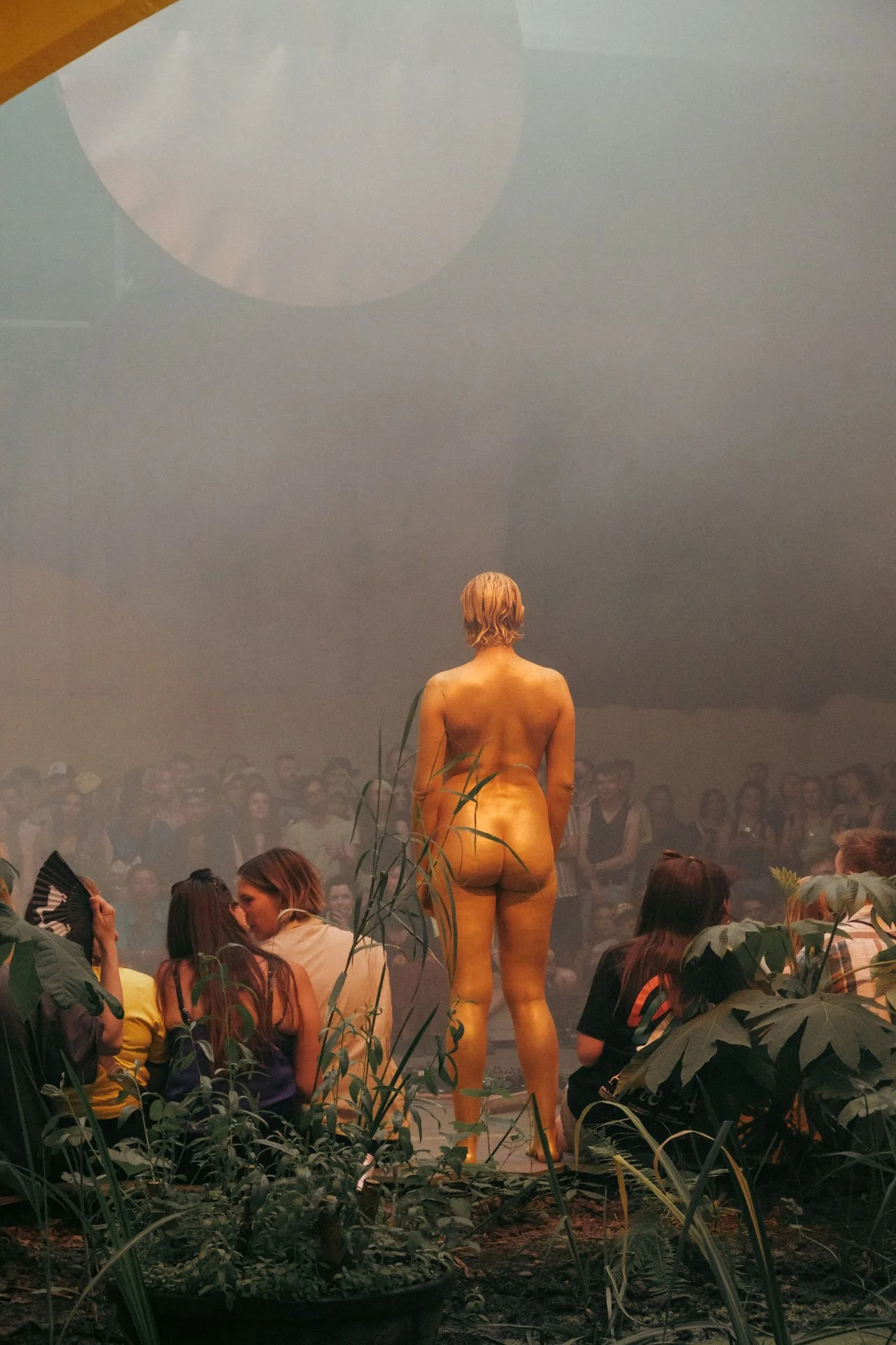 Personne nue vue de dos, assise dans un environnement artistique ou théâtral, avec un public assis devant, entourée de plantes.