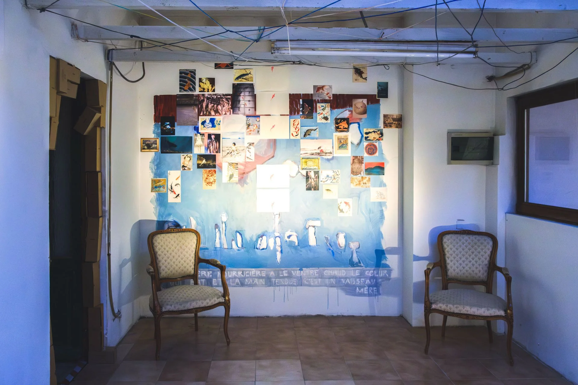 Une salle d'exposition avec deux fauteuils en bois et tissu beige, un mur orné d'une mosaïque d'images et de peintures abstraites, et une fenêtre sur le côté droit