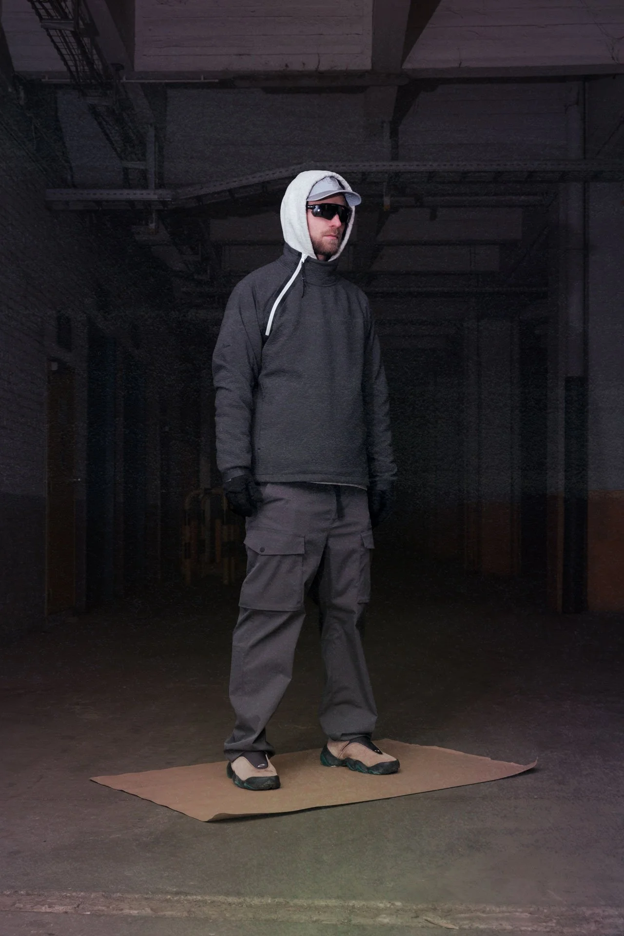 Un homme portant des vêtements de plein air noirs, un chapeau gris, des lunettes de soleil et des gants, debout sur un tapis en carton dans un espace sombre, probablement un entrepôt ou un atelier.
