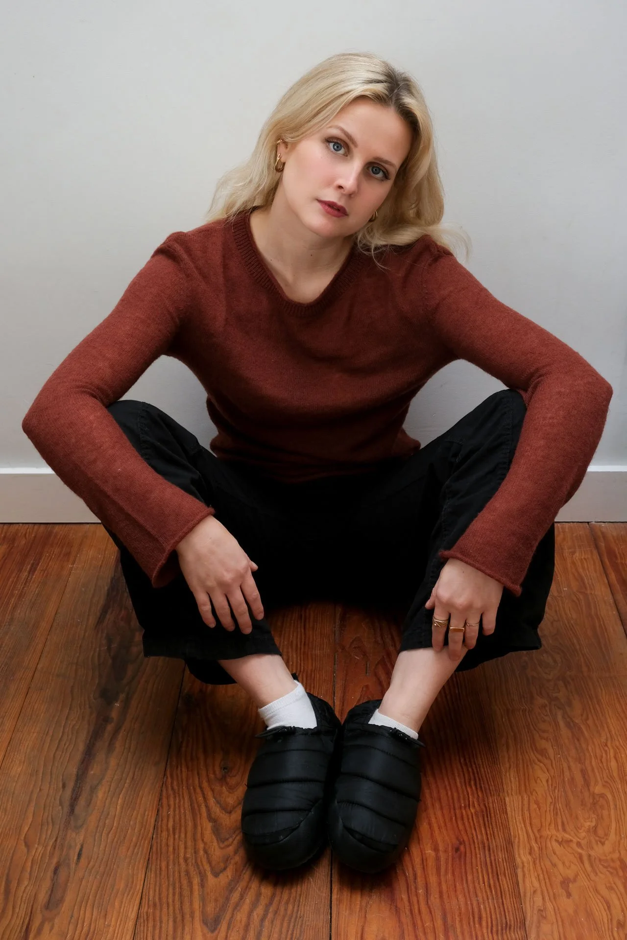 Une femme blonde en pull marron, assise par terre sur un sol en bois, en position de squat, regardant vers la caméra, avec un fond blanc.