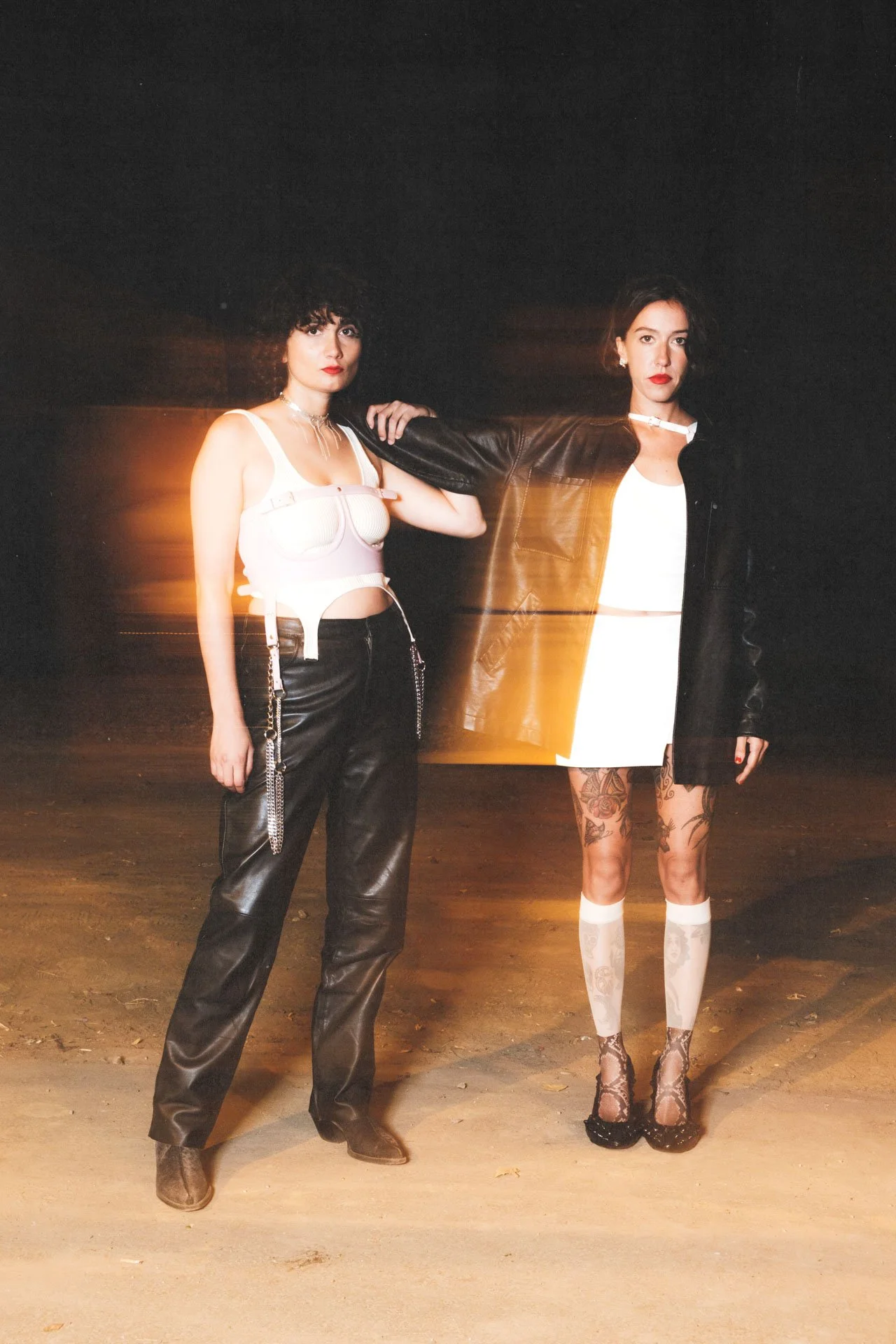 Deux femmes posent dans un environnement sombre. La femme à gauche porte un haut blanc et un pantalon en cuir, tandis que celle à droite porte une veste en cuir noir et une jupe blanche, avec des tatouages visibles sur ses jambes.
