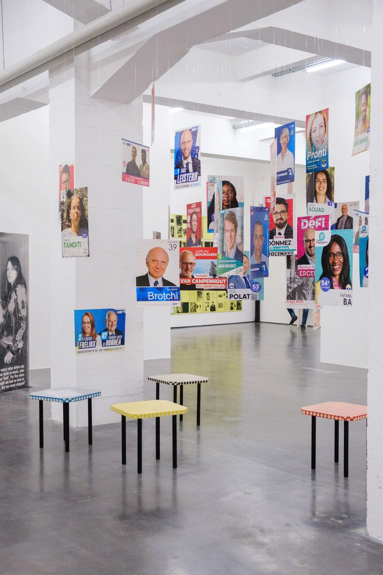 Exposition avec affiches politiques suspendues au plafond dans une galerie d'art, avec des sièges colorés en avant-plan.