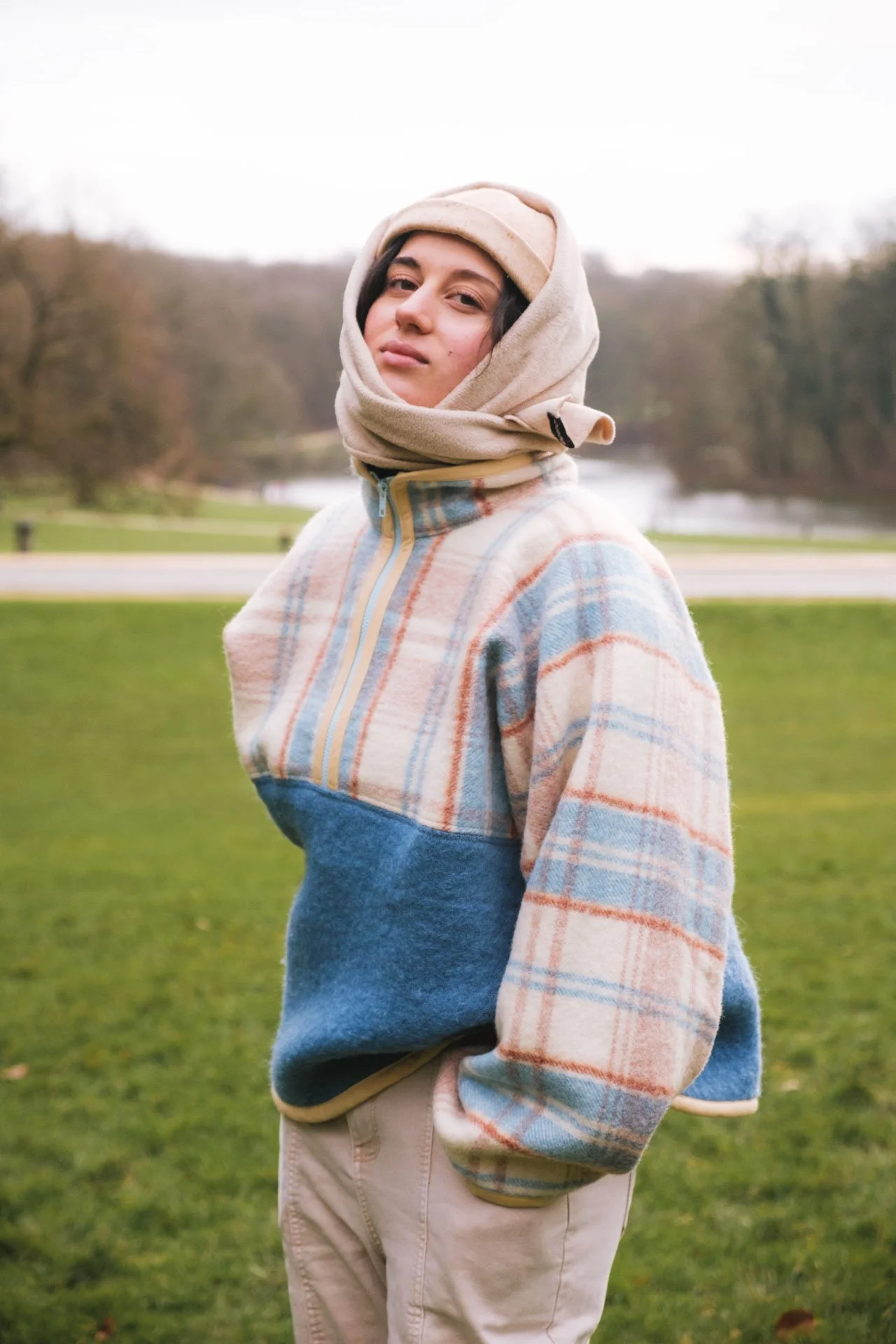 Jeune femme portant un bonnet, une écharpe et un pull plaid dans un parc verdoyant avec un lac en arrière-plan.